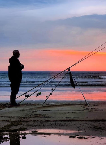 Person fishing on a beach at sunset with fishing rods from the angling hub.